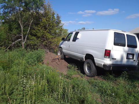 1999 Ford E250 - A white van is parked on a dirt hill surrounded by greenery under a blue sky with clouds.