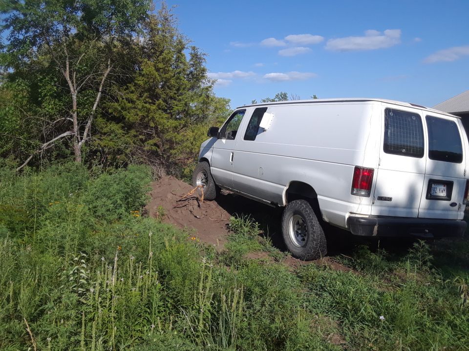 White van with off-road tires on a dirt incline, trees and grass around. License plate visible