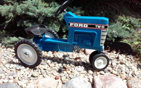 1994 Ford Bronco - A blue Ford TW-5 pedal tractor sits on a rocky surface next to some evergreen bushes.