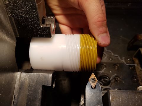 1978 Ford Bronco - A hand holds a white and yellow threaded pipe fitting near a metal lathe cutting tool. The fitting is being held in the chuck of the lathe.