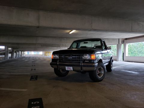 1988 Ford Bronco - A black 1988 Ford Bronco is parked in a concrete parking garage. The front 3/4 view shows the vehicle's grille, headlights, and raised susp…