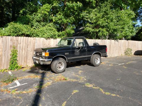 1988 Ford Bronco - A black 1988 Ford Bronco is parked in a lot next to a wooden fence. The vehicle has off-road tires and a spare tire mounted in the bed.