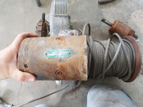1978 Ford Bronco - A person's hand holds a rusty electric winch motor with a spool of cable wrapped around it. A green and white label on the motor reads "PRE…