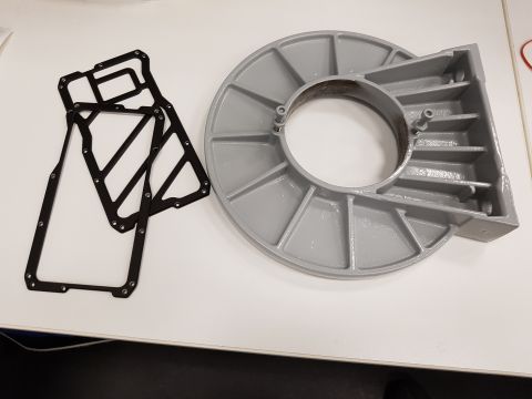 1978 Ford Bronco - Two black gaskets and a gray metal part with a circular opening and radiating fins are on a white surface.