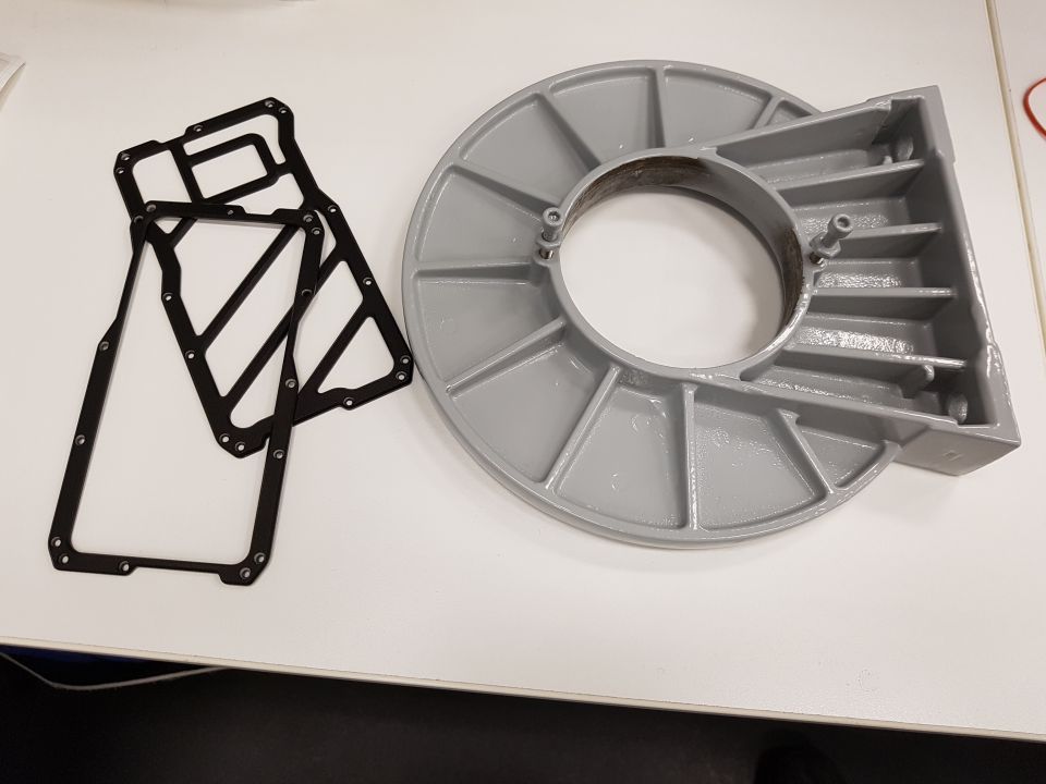 Two black gaskets and a gray metal part with a circular opening and fins.