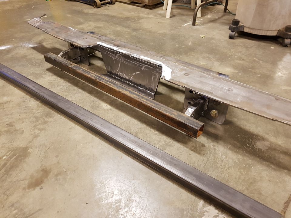 Metal frame pieces on a concrete floor, one long square tube and a complex welded assembly