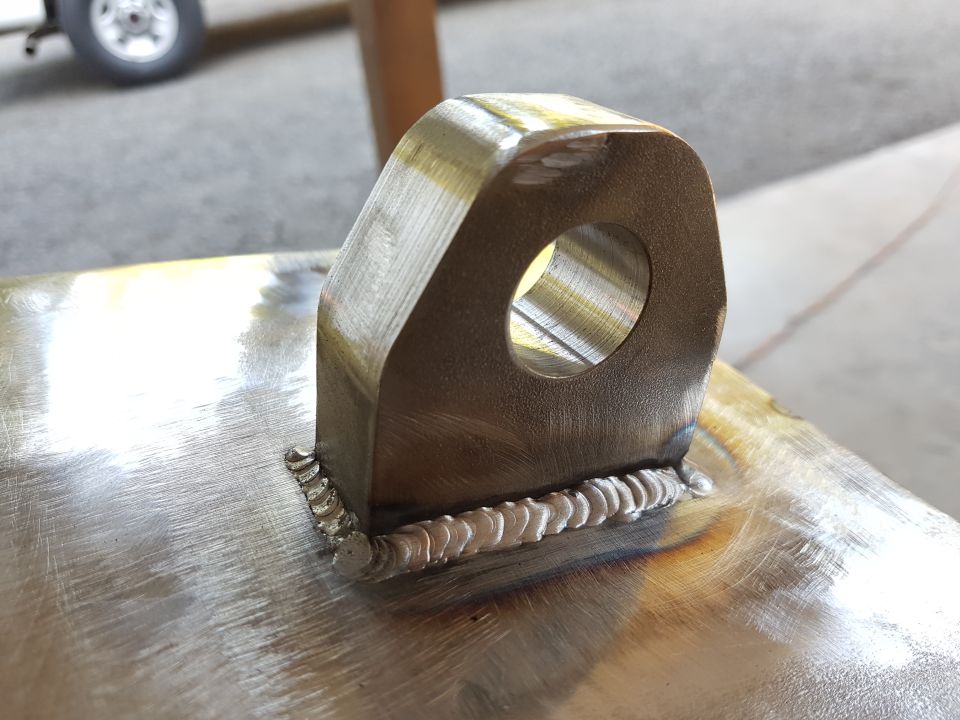 Close-up of a metal bracket with a hole, attached by welds. A wheel is visible in the blurred background