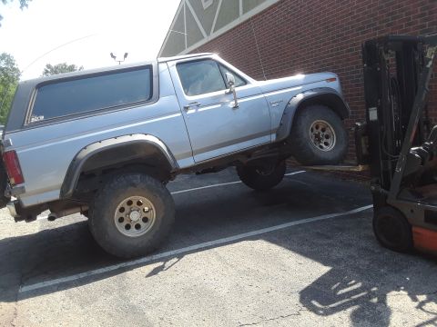 1985 Ford Bronco - A forklift is lifting the rear of a light blue Ford Bronco, which is parked in a lot next to a brick building.