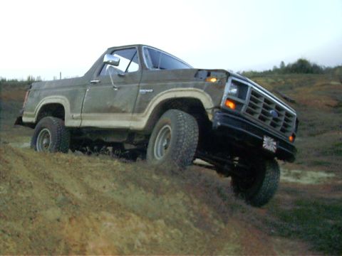 1985 Ford Bronco - A brown Ford Bronco navigates a muddy incline, its front wheels partially submerged.