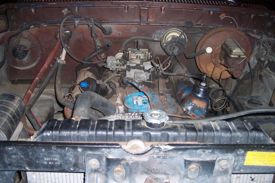Engine bay showing carburetor, radiator, brake booster, and hoses. Some text visible on radiator