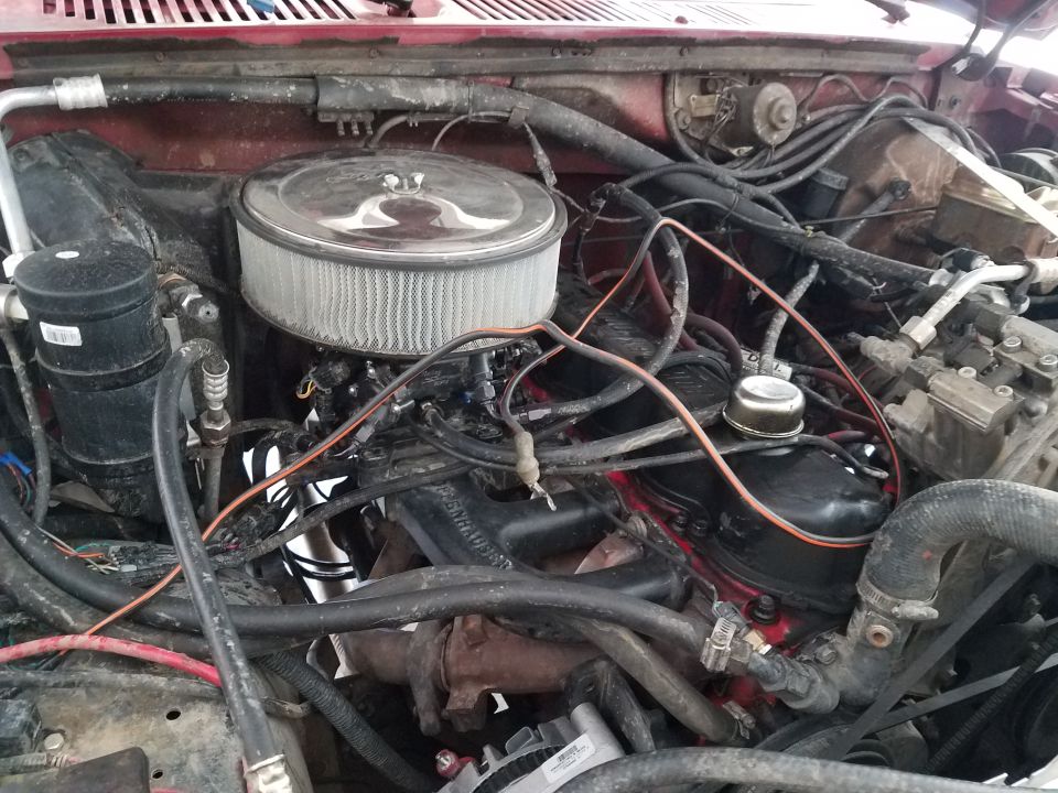 Engine bay of a red vehicle, showing a V8 engine with an aftermarket air filter and various hoses and wires. The condition appears used