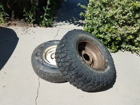 1985 Ford Bronco - Two BFGoodrich Mud-Terrain tires and rims are shown resting on a concrete surface next to some greenery.