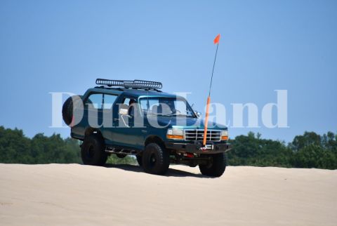 1996 Ford Bronco - This 1996 Ford Bronco is seen from a 3/4 front view, equipped with a roof rack and spare tire, traversing sand dunes under a clear blue sky.