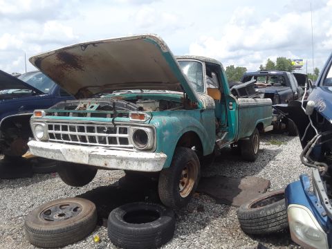 1994 Ford Bronco - A teal 1965 Ford F-Series truck sits in a junkyard with its hood open and tires removed.