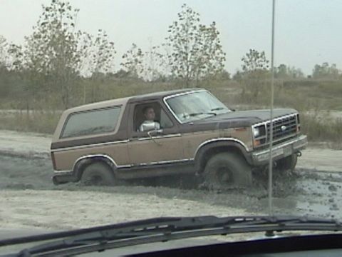 1995 Ford Bronco - A two-tone Ford Bronco is partially submerged in mud, with trees and an overcast sky in the background.