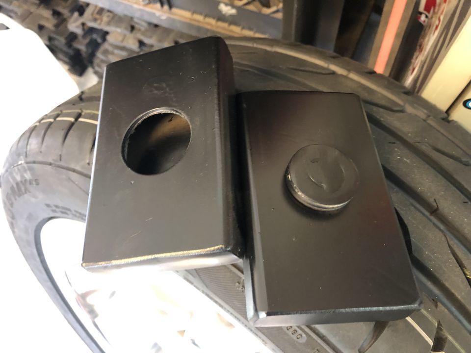 Two black metal blocks, one with a hole, sit on a tire. One block has a bolt head visible