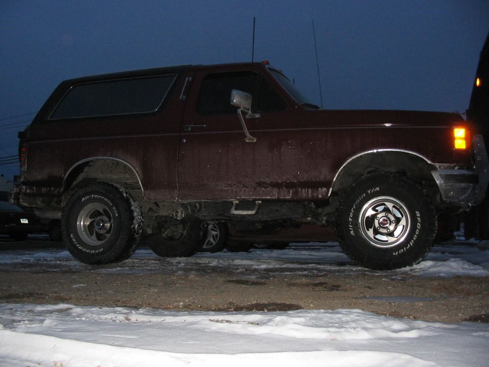 Side view of a lifted burgundy Ford Bronco on BFGoodrich All-Terrain tires, parked on snowy muddy ground at dusk