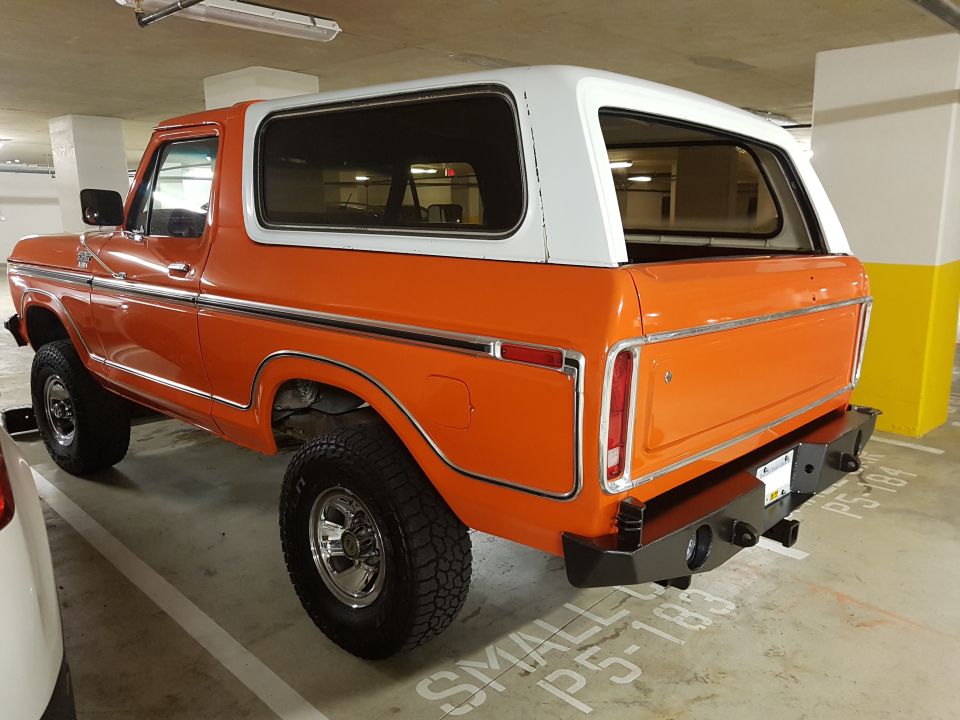 Orange Ford Bronco parked in a garage, showing its rear and side profile. It has a white top and chrome trim