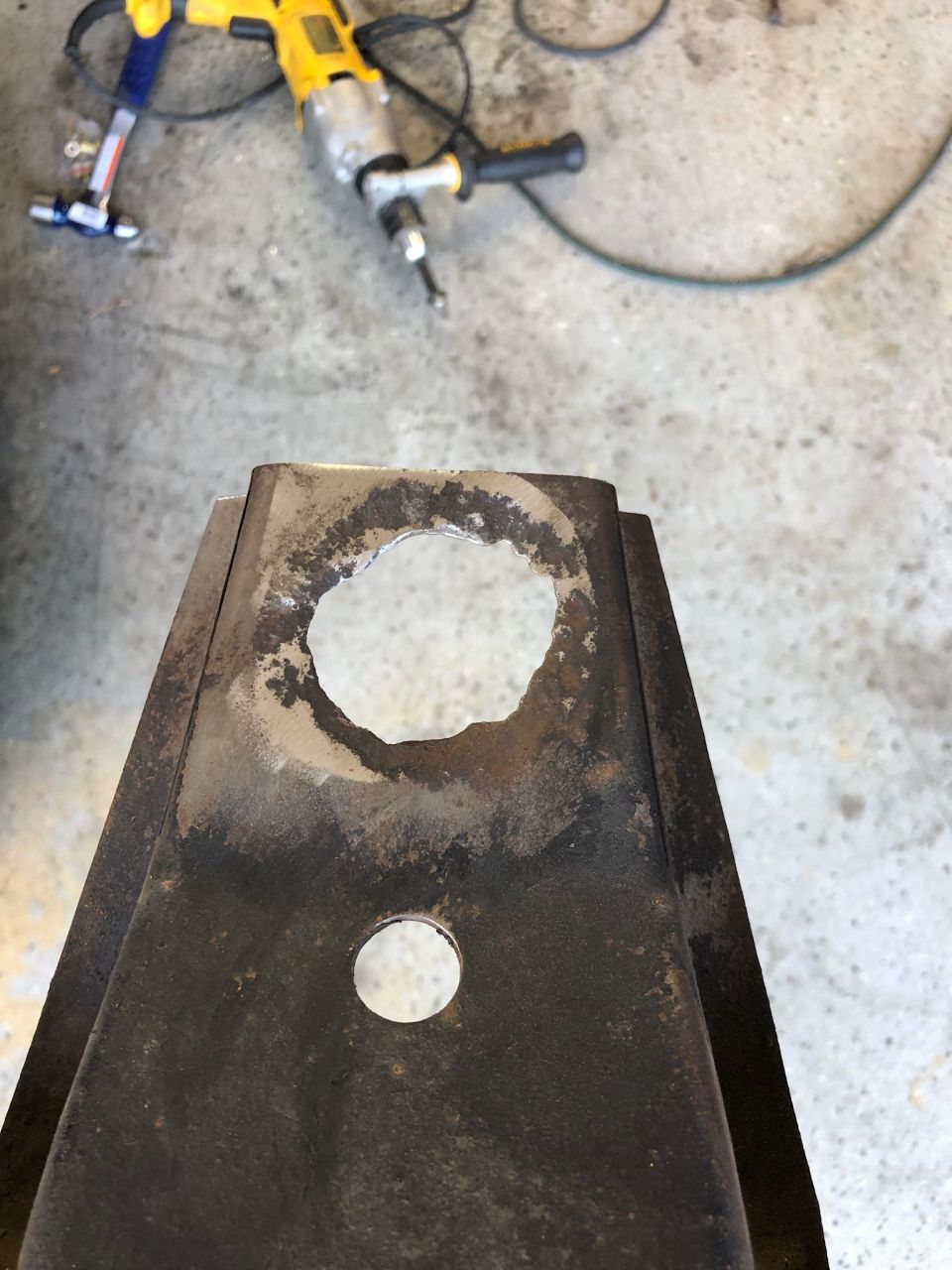 Close-up of a rusty metal part with a large hole drilled through it. A yellow drill is visible in the background