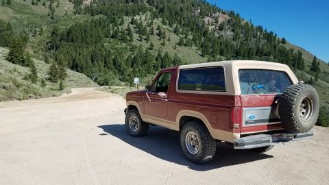1985 Ford Bronco - This Ford Bronco is parked on a dirt road with a mountainous, forested landscape behind it under a clear blue sky.