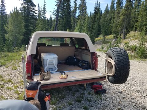 1985 Ford Bronco - A Ford Bronco is parked on a gravel road in a wooded area, its tailgate open to reveal camping gear.