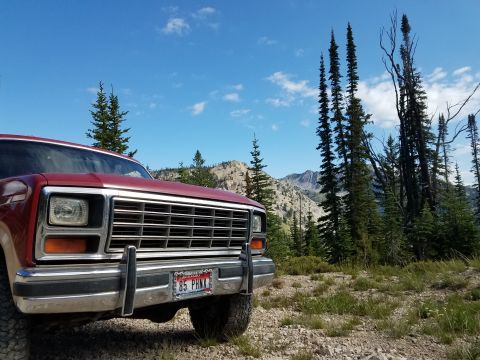 1985 Ford Bronco - A red Ford Bronco is parked on a gravel road with mountains and trees in the distance under a blue sky.