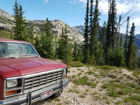 1985 Ford Bronco - 1985 Ford Bronco parked on a mountain road with trees and rocky hills under a blue sky.
