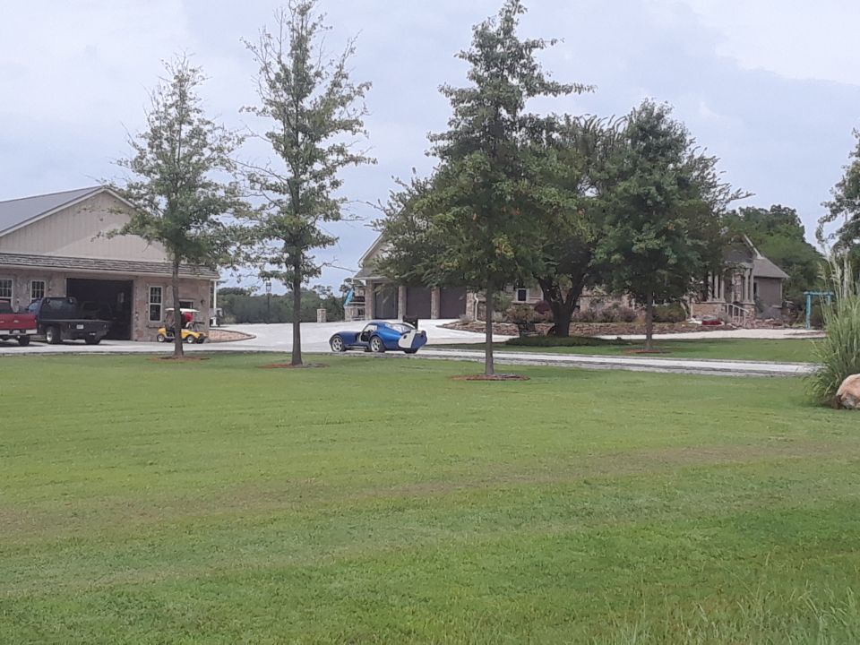 Blue sports car with doors open parked in driveway of large house. Two trucks are parked in an open garage