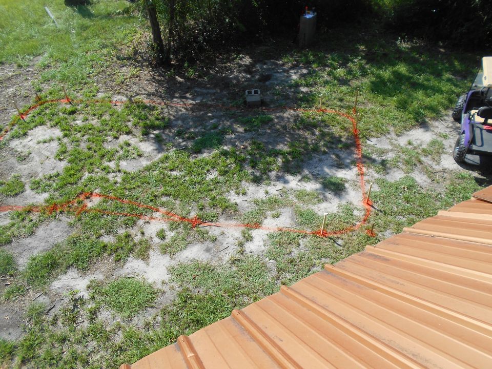 orange marking tape outlines a construction area on a grassy lot, with a cinder block and some stakes visible. A purple ATV is parked to the right