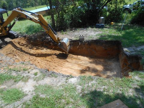 1992 Ford Bronco - An excavator is actively digging a deep hole in a yard, with trees and a building in the background.