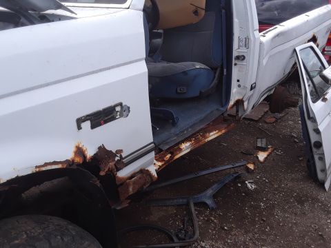 1994 Ford Bronco - A heavily rusted white pickup truck sits in a junkyard, revealing its worn interior and damaged exterior.