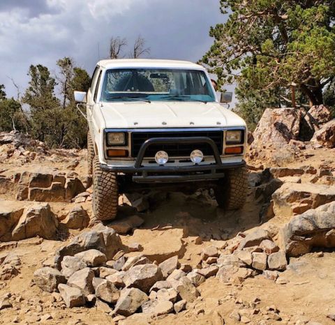1981 Ford Bronco - White Ford Bronco tackling a challenging rocky off-road path under a cloudy sky.