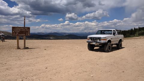 1995 Ford Bronco - A white Ford Bronco is parked on a dirt road with a signpost and mountains in the distance.