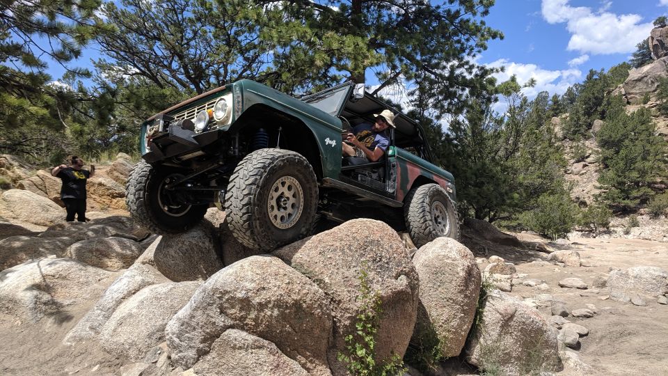 Green Ford Bronco with large tires navigating a rocky trail. Driver wears a hat. Another person stands on rocks in background