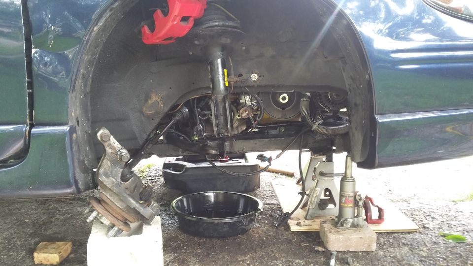 Car's front suspension disassembled with red brake calipers, engine components visible, and oil drain pans on the ground