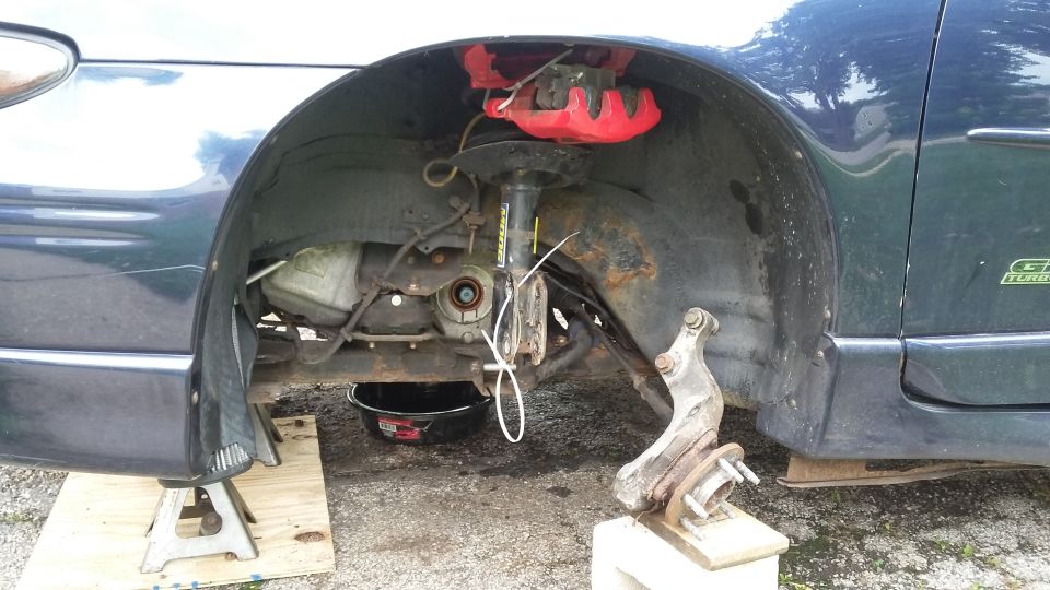 Dark blue car's front wheel assembly removed, with red brake caliper and oil pan visible