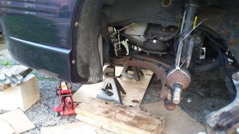 Front corner of a vehicle raised on a floor jack and stands showing rusty front suspension