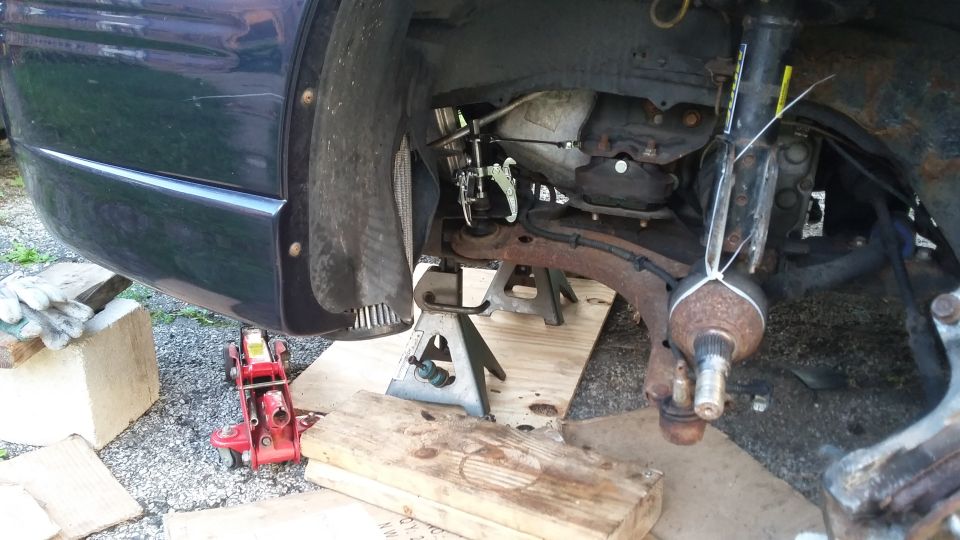 Front corner of a vehicle raised on a floor jack and stands showing rusty front suspension
