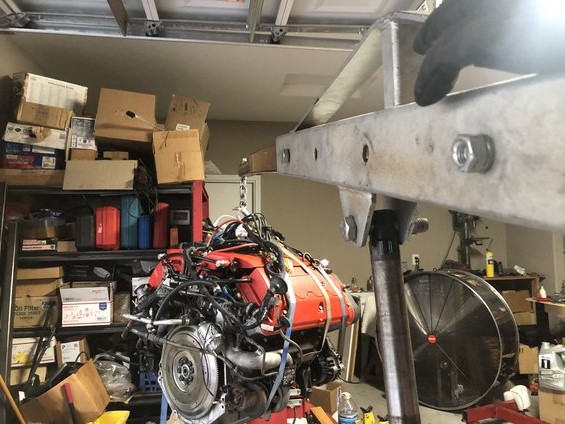 Engine assembly on a red stand, suspended by a metal hoist in a cluttered garage