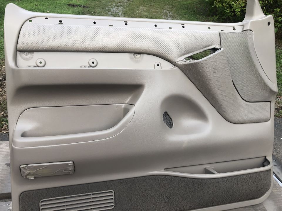 Car door panel with storage pocket, speaker grille, and torn fabric trim. Tape covers a small rectangular handle