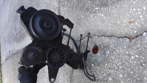 1994 Ford Bronco - A detached power steering pump assembly with multiple pulleys sits on a cracked concrete surface.