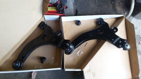 Two black control arms with ball joints and mounting hardware in cardboard boxes