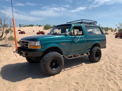 1996 Ford Bronco - Teal 1996 Ford Bronco with off-road modifications parked on sand dunes under a blue sky.