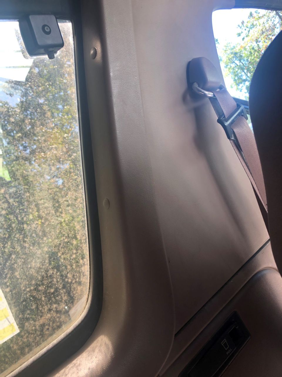 Tan interior pillar with a brown seatbelt and a window showing trees. A Supermotors.net watermark is visible
