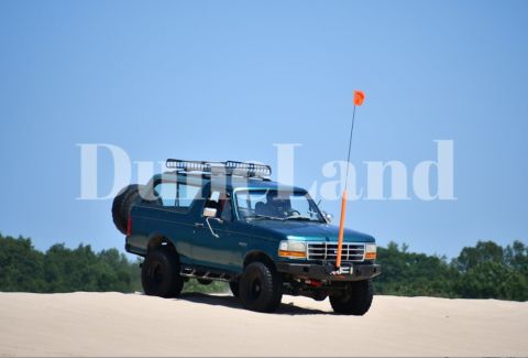 1996 Ford Bronco - A teal 1996 Ford Bronco is shown driving on a sandy terrain under a clear blue sky.