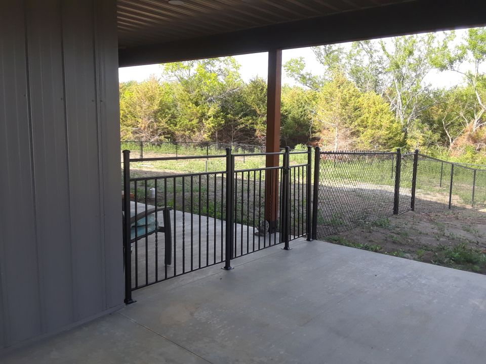 Backyard patio area with a chain-link fence and gate, leading to trees and brush
