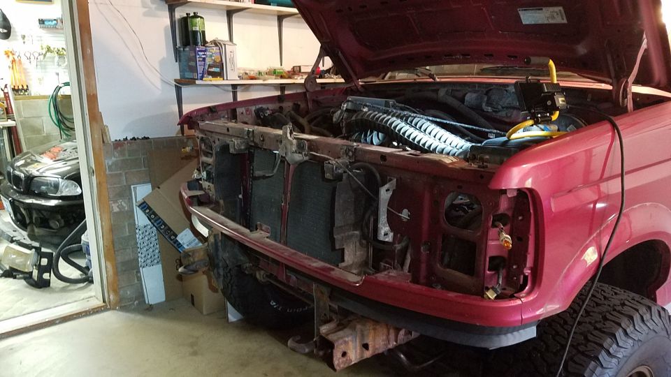 Red Ford Bronco with hood open, front end disassembled, showing radiator and engine components. Visible tire and fender