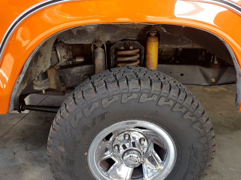 1978 Ford Bronco - Close-up of the front wheel well and suspension of an orange 1978 Ford Bronco, a chrome rim and off-road tire.