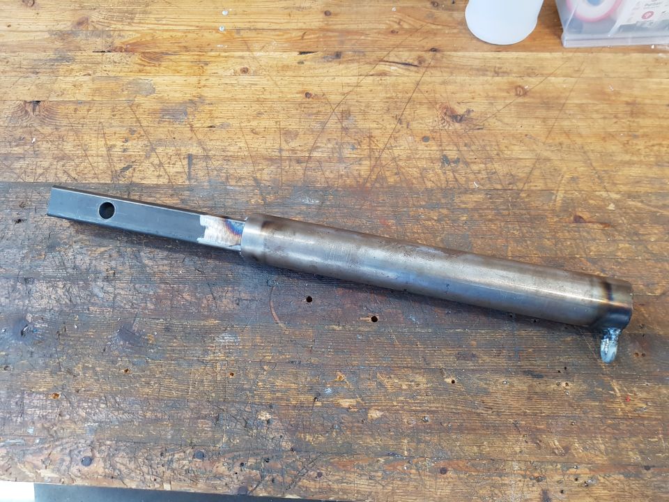 Custom fabricated metal part with a square end and a round tube section, resting on a wooden workbench