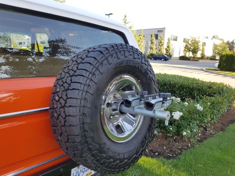 1978 Ford Bronco - A spare tire is mounted on the rear of an orange SUV, with a building and trees in the background.